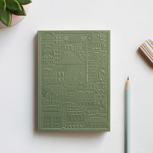 green debossed notebook with city buildings, bridges and river on grey table with pencil, stripy eraser and plant in top left corner