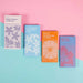 four rectangle packets against a pink and blue background. One Packet is white, one blue, one orange and on lilac 