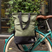 GREEN AND BLACK FABRIC BAG WITH 3 STRAPS ATTACHED TO A BLUE BICYCLE WITH BROWN  SADDLE IN FRONT OF A GREEN BUSH