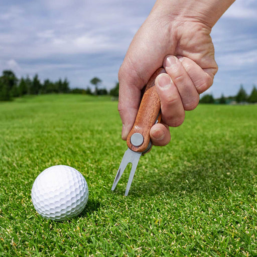white golf ball on green grass with a hand holding a wooden and metal forked tool to the grass with blue sky in the background 
