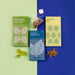 3 rectangle boxes, one green, one blue and one yellow in front of a mint and blue background. With teabag, green leaves and seeds 