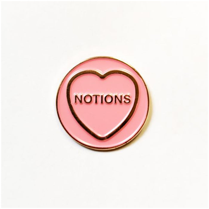 pink circular enamels pin with gold circumference, a gold heart and text in the heart that reads 'notions' in front of a white background