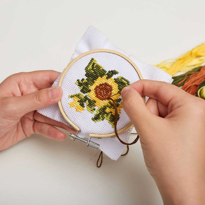 pair of hands holding a needle and thread and embroidering a sunflower in yellow, brown and green thread