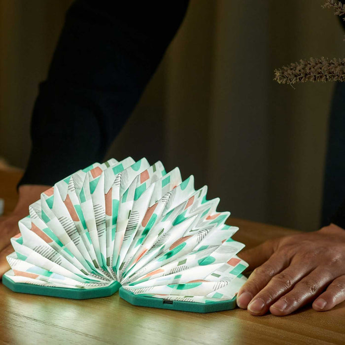 accordion shaped light open in a half circle with green base and pink, green and white pattern cover. Light in on a wooden table between a pair of hands