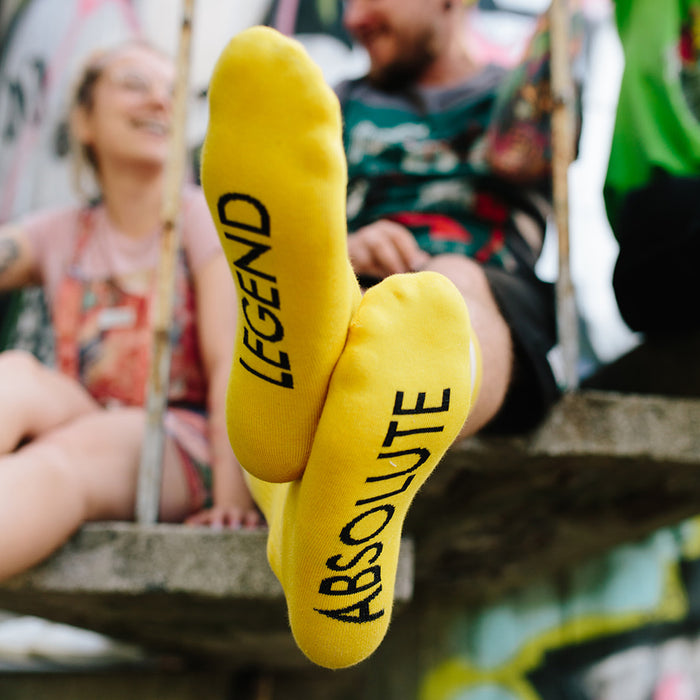 yellow socks on males with absolute legend written on sole in black capital letters, you can only see the sole of his feet in focus.. Photo was taken to focus in on socks, in background is blurred out two people sitting on a bench chatting.