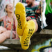 yellow socks on males with absolute legend written on sole in black capital letters, you can only see the sole of his feet in focus.. Photo was taken to focus in on socks, in background is blurred out two people sitting on a bench chatting.