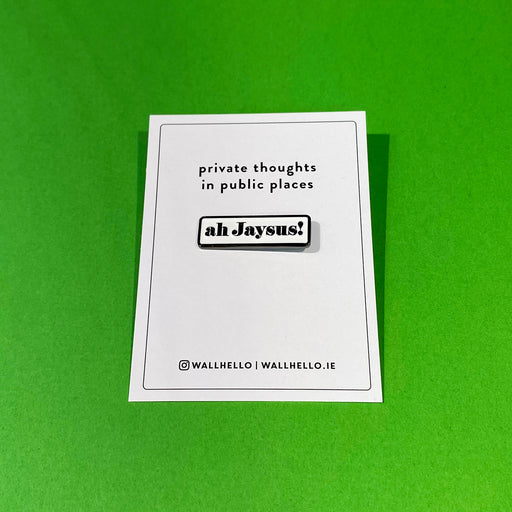 white backing card with black text and white enamel pin with 'ah jaysus!' in black on a green background