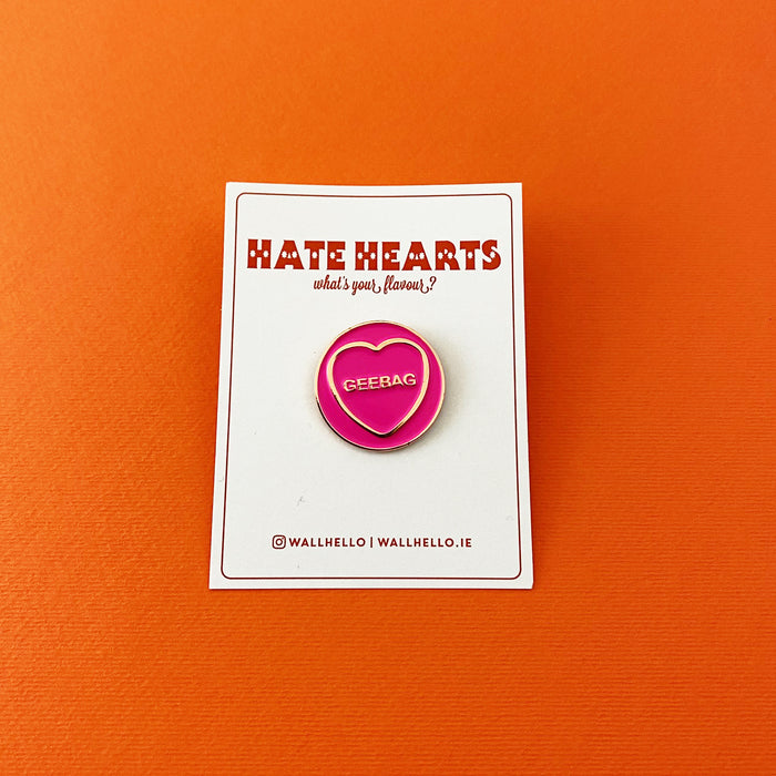 circular pink enamel pin with gold circumference, a gold heart and text in the heart that reads 'geebag' on a white backing card in front of an orange background