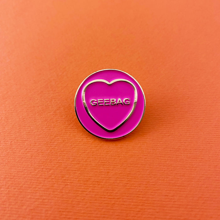 circular pink enamel pin with gold circumference, a cold heart and text in the heart that reads 'geebag' on an orange background