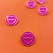 5 circular pink enamel pins with gold circumference, a cold heart and text in the heart that reads 'geebag' on an orange background