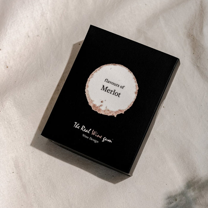 A black rectangular product box with a circular white and beige label reading 'flavours of Merlot' and smaller text 'The Real Wine Gum Wine Design,' photographed on a light fabric surface.