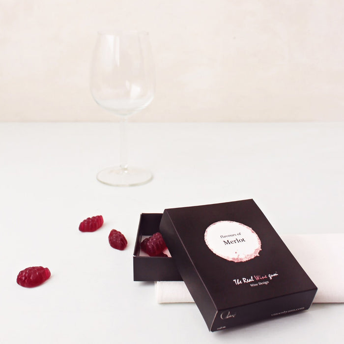 A black rectangular product box with a circular white and beige label reading 'flavours of Merlot' photographed on a light fabric surface next to an empty wine glass and some red gummies.