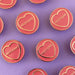 pink circular enamels pins with gold circumference, a gold heart and text in the heart that reads 'notions' in front of a purple background