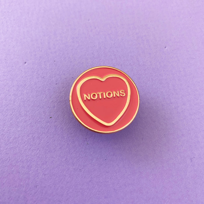 pink circular enamels pin with gold circumference, a gold heart and text in the heart that reads 'notions' in front of a purple background