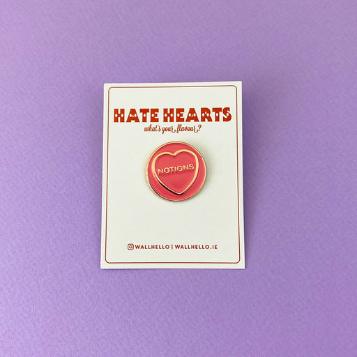 pink circular enamels pin with gold circumference, a gold heart and text in the heart that reads 'notions' on a white backing card  in front of a purple background