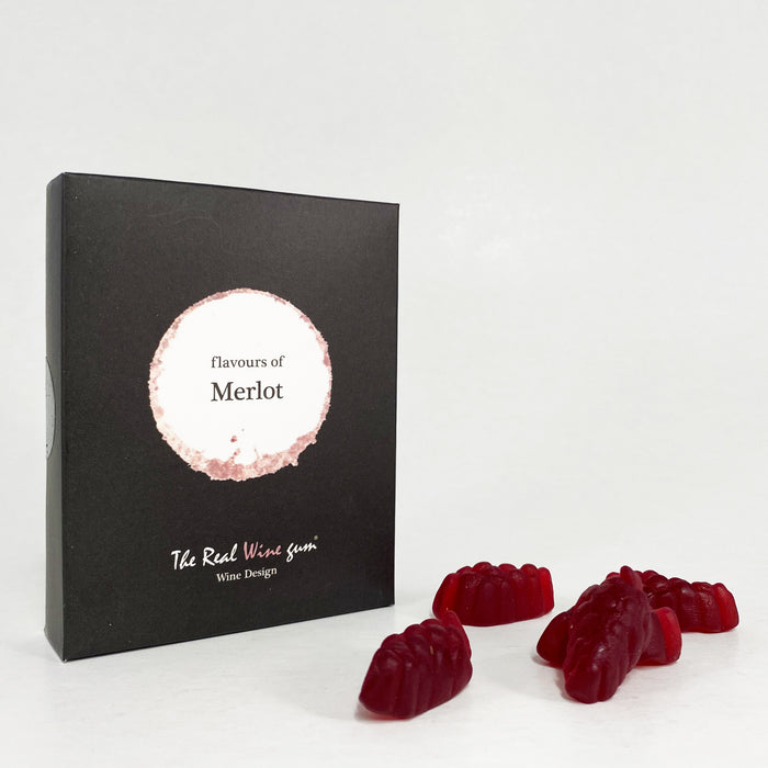 A black rectangular product box with a circular white and beige label reading 'flavours of Merlot' and smaller text 'The Real Wine Gum Wine Design,' photographed on a light fabric surface next to some red gummies.