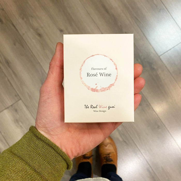 A hand holding a cream rectangle box with "Flavours of Rose Wine" text against a wooden shop floor.