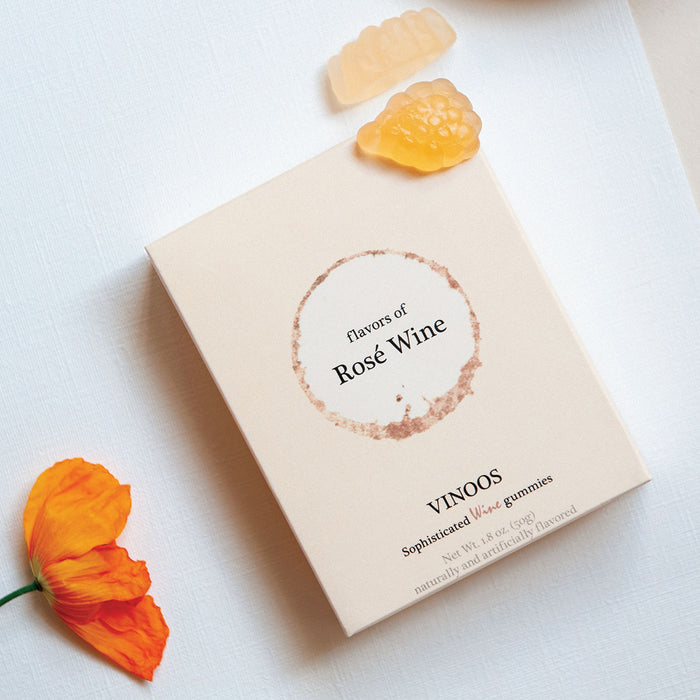 A cream rectangular product box with a circular white and sand coloured label reading 'flavours of Rose Wine' photographed on a light fabric surface next to an orange flower and two yellow gummies.