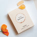 A cream rectangular product box with a circular white and sand coloured label reading 'flavours of Rose Wine' photographed on a light fabric surface next to an orange flower and two yellow gummies.