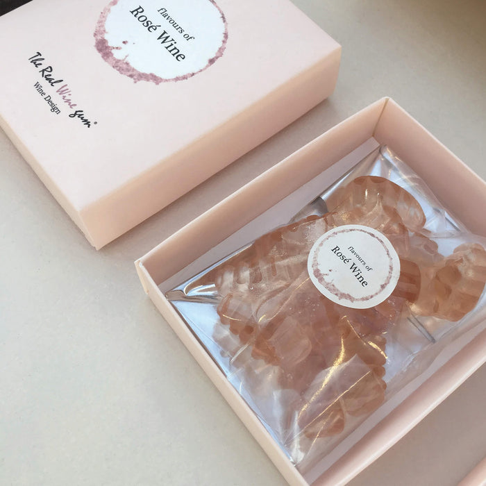 An open cream rectangular product box with a circular white and beige label reading 'flavours of Rose Wine' on the front and a plastic bag of pink coloured gummies inside photographed on a light fabric surface.