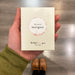 A hand holding a light green rectangle box with "Flavours of Sauvignon" text against a wooden shop floor.