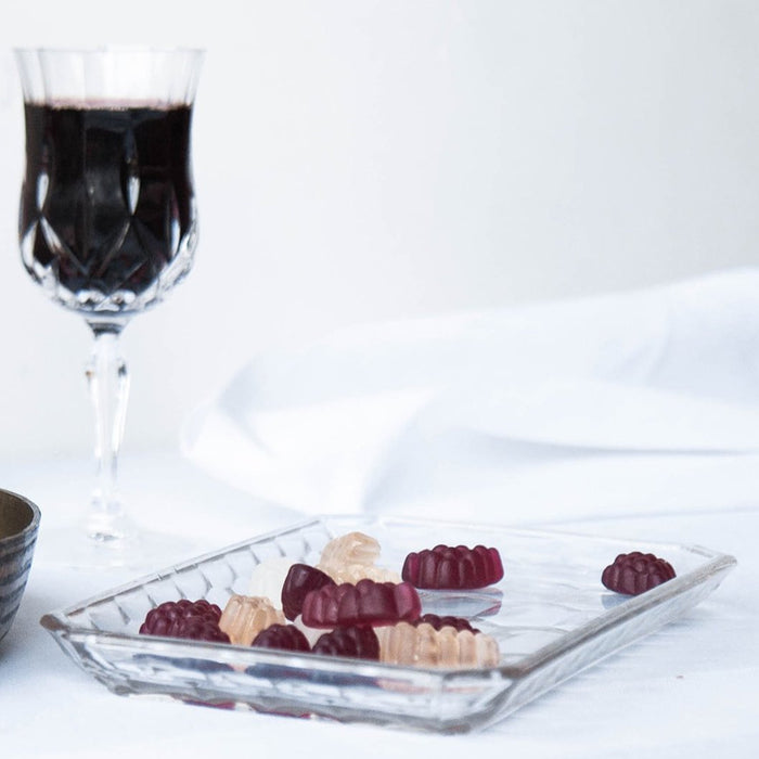 A glass dish of assorted gummies with a glass of red wine in the background against a white backdrop.