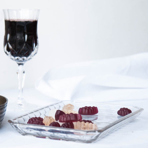 A glass dish of assorted gummies with a glass of red wine in the background against a white backdrop.