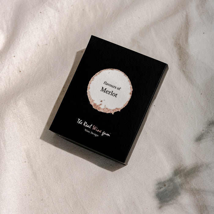 A black rectangular product box with a circular white and beige label reading 'flavours of Merlot' and smaller text 'The Real Wine Gum Wine Design,' photographed on a light fabric surface.