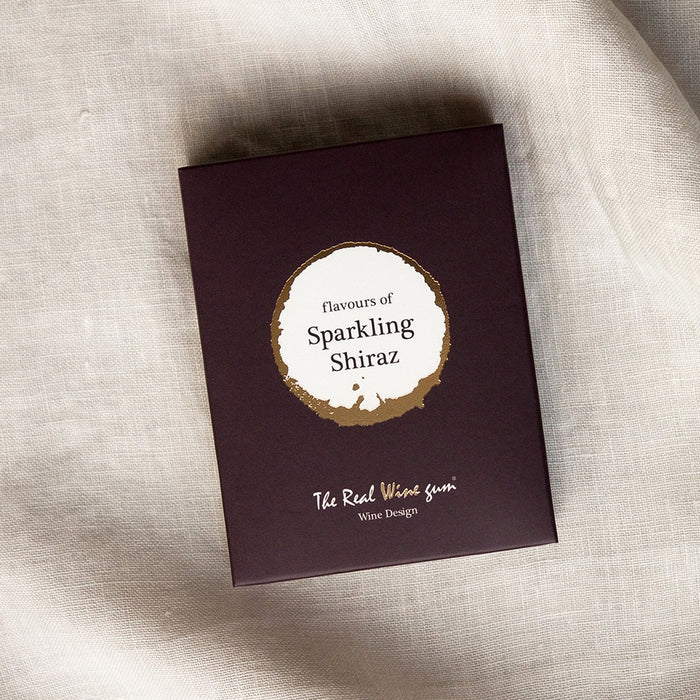 A wine coloured rectangular product box with a circular white and beige label reading 'flavours of Sparkling Shiraz' and smaller text 'The Real Wine Gum Wine Design,' photographed on a light fabric surface.