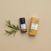Rosemary essential oil blue bottle beside an orange product box with an illustration of a rosemary leaf on front with rosemary sprig to he left against a pink background