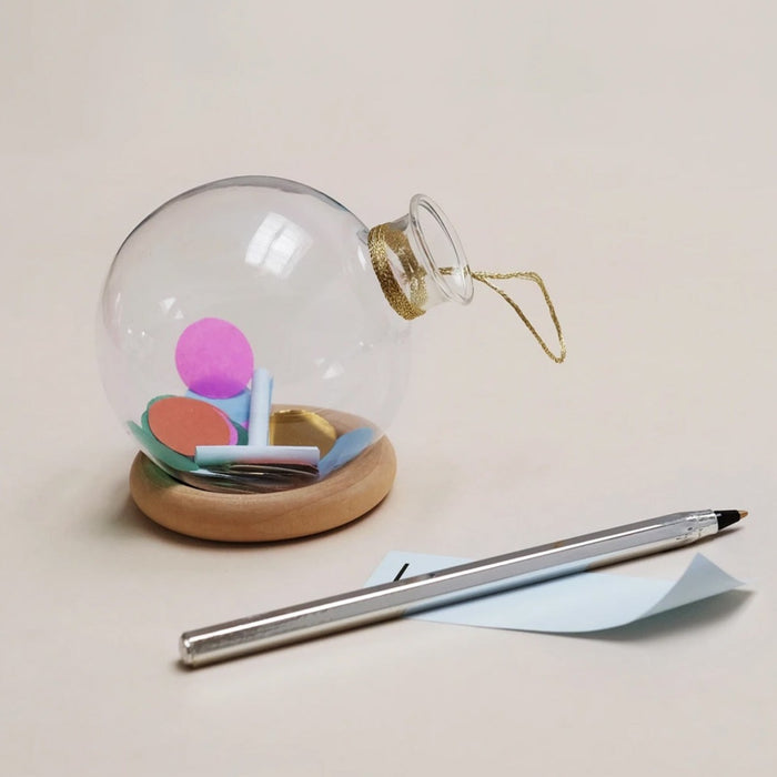 glass globe on a wooden ring stand with confetti  and scrolls of paper inside beside a pen